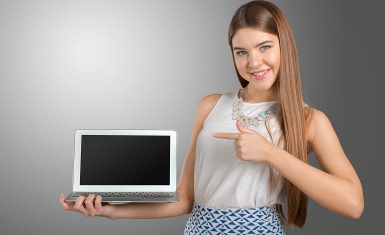 A smiling young woman holding and pointing at a sleek, silver laptop with a blank screen, showcasing the best laptop for work and play.HP Dell and Lenovo Laptops of 2025