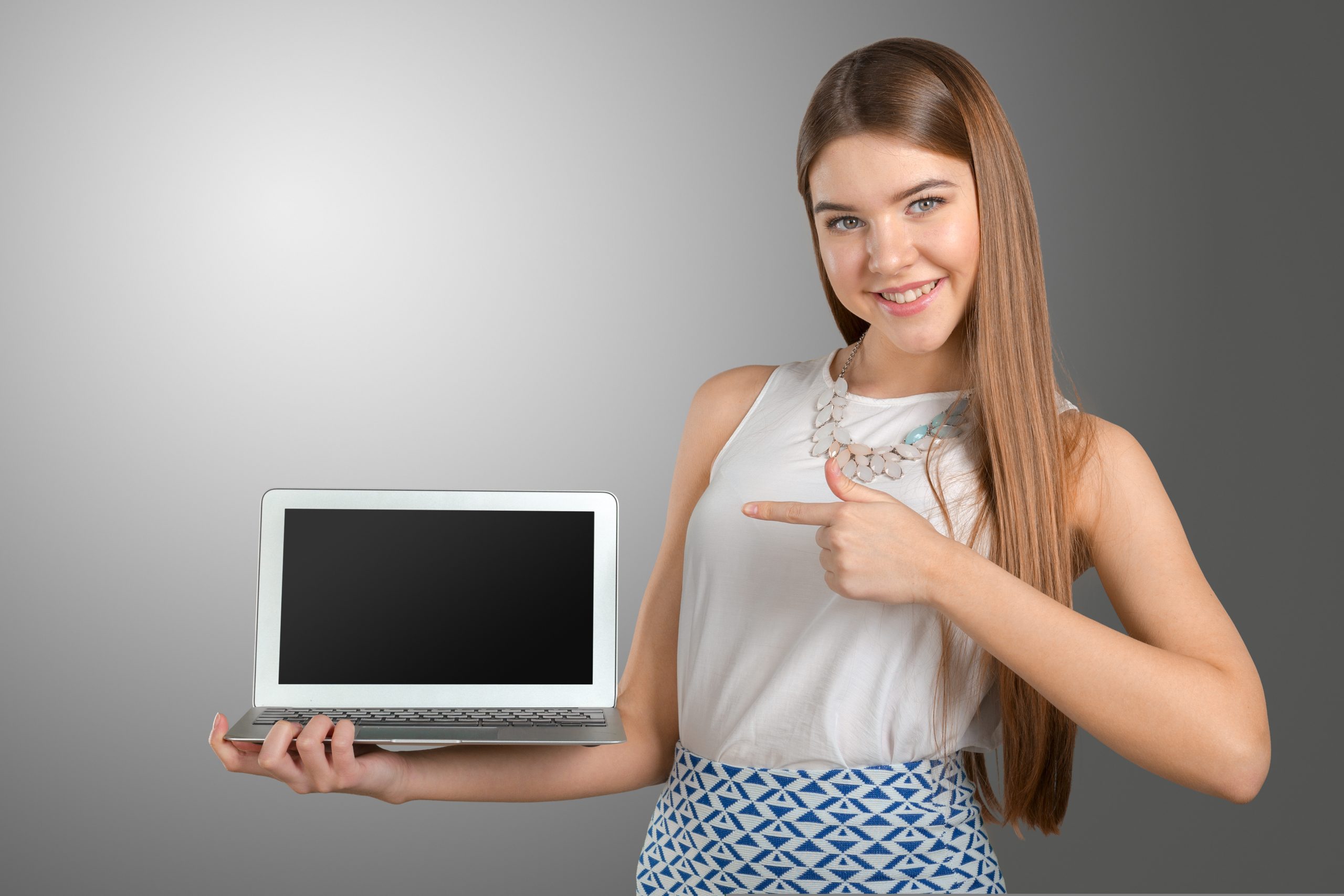 A smiling young woman holding and pointing at a sleek, silver laptop with a blank screen, showcasing the best laptop for work and play.HP Dell and Lenovo Laptops of 2025