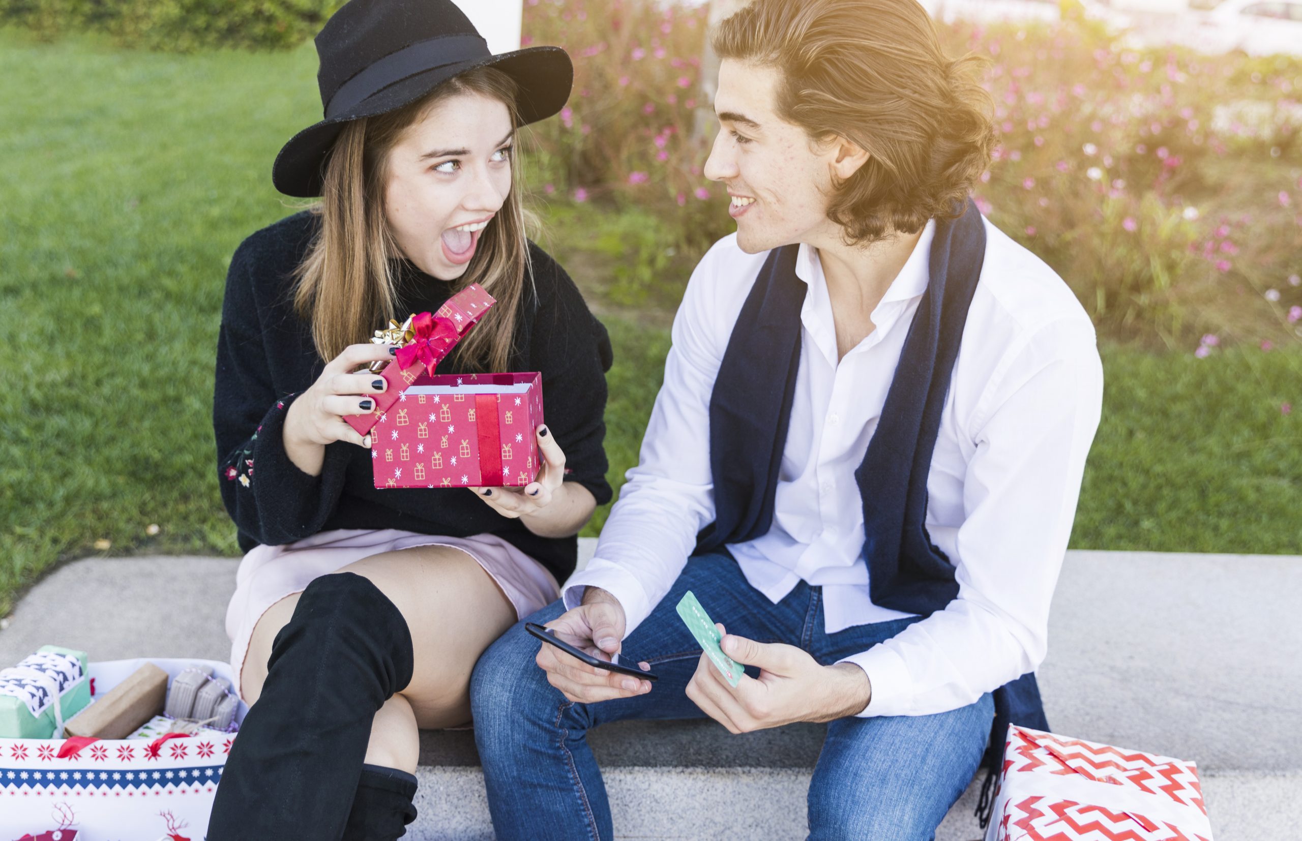 A young woman excitedly opening a red gift box while sitting outdoors with her male friend, highlighting the joy of exchanging thoughtful presents