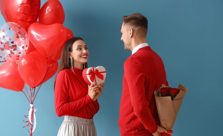 A young couple in red outfits celebrating Valentine's Day. The woman holds a heart-shaped gift box while the man hides a bouquet of roses behind his back. Red heart-shaped balloons are in the background.