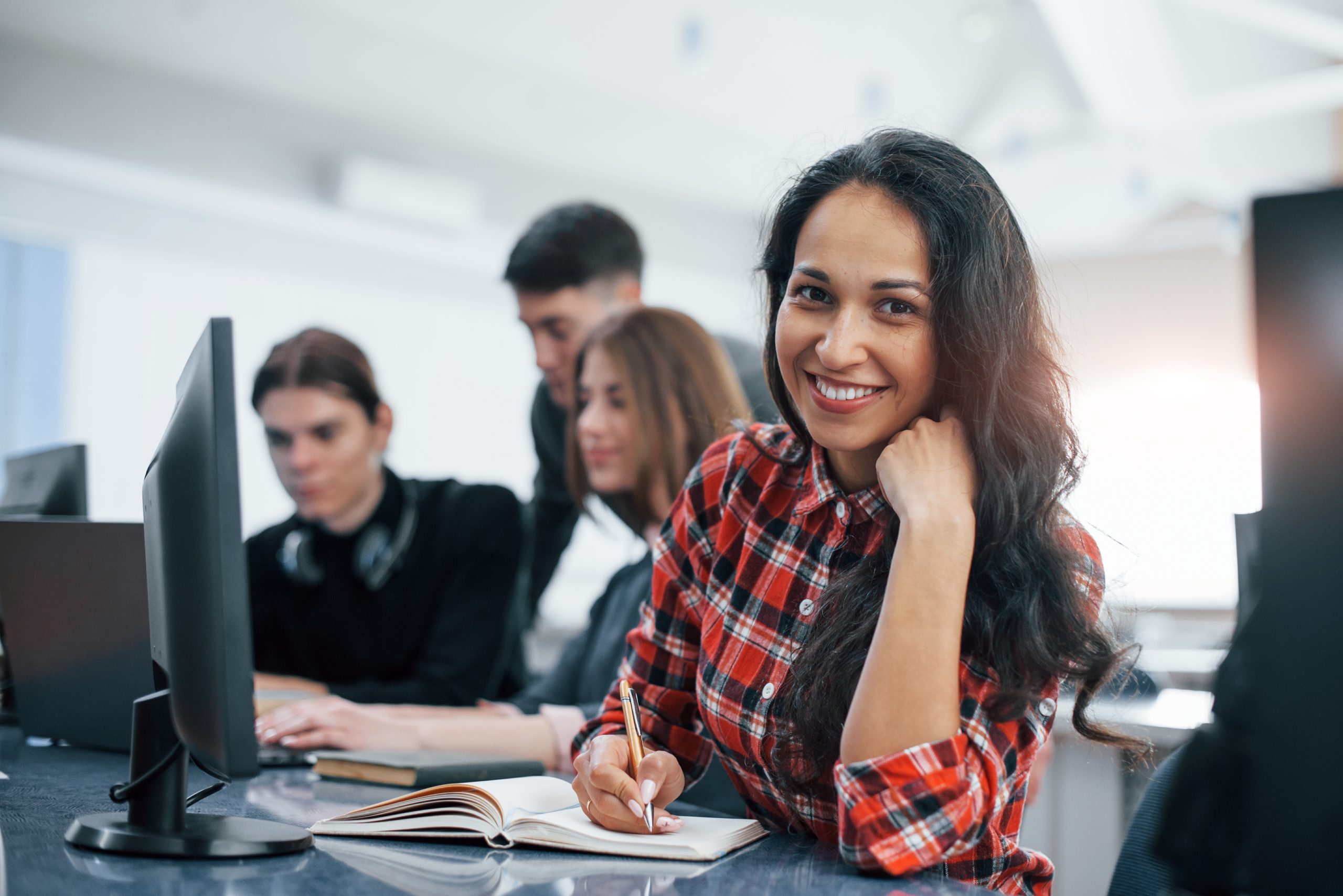 A smiling young woman in a red checkered shirt sitting in a modern classroom, writing in a notebook, with classmates working on computers in the background.