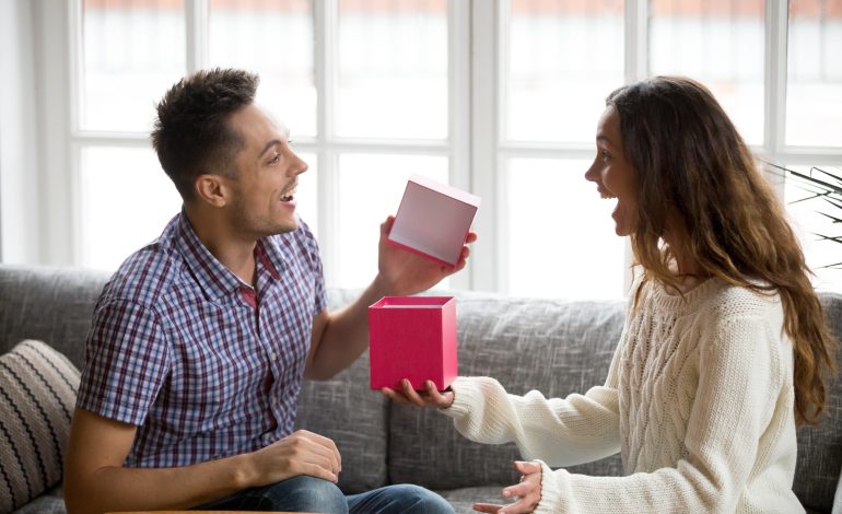 A young man excitedly opens a pink gift box while sitting on a couch with a smiling woman, both expressing joy and surprise—perfect for inspiring DIY Gift Ideas for Loved Ones.